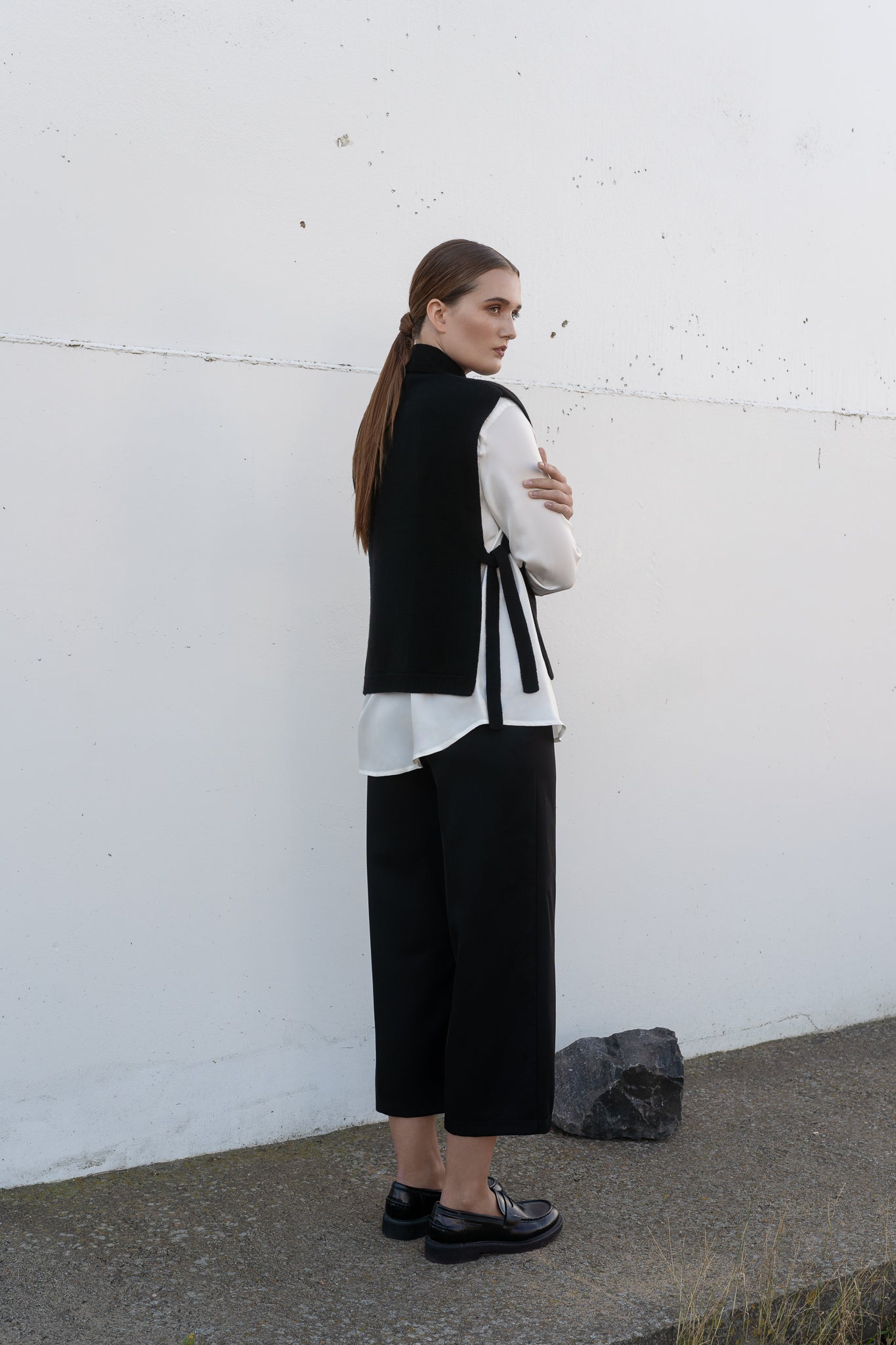 Woman wearing a black vest over a white shirt with black pants against a white wall.