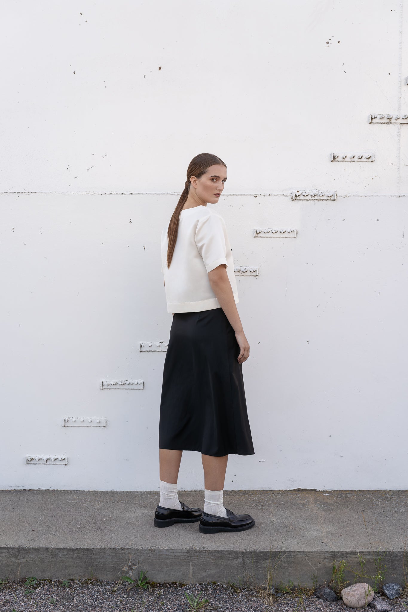 model wearing black wool midi skirt and white silk top agains industrial back wall.