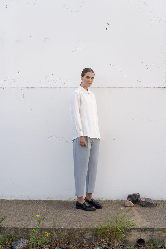 A model wearing white silk shirt and greyish blue trousers against industrial background.