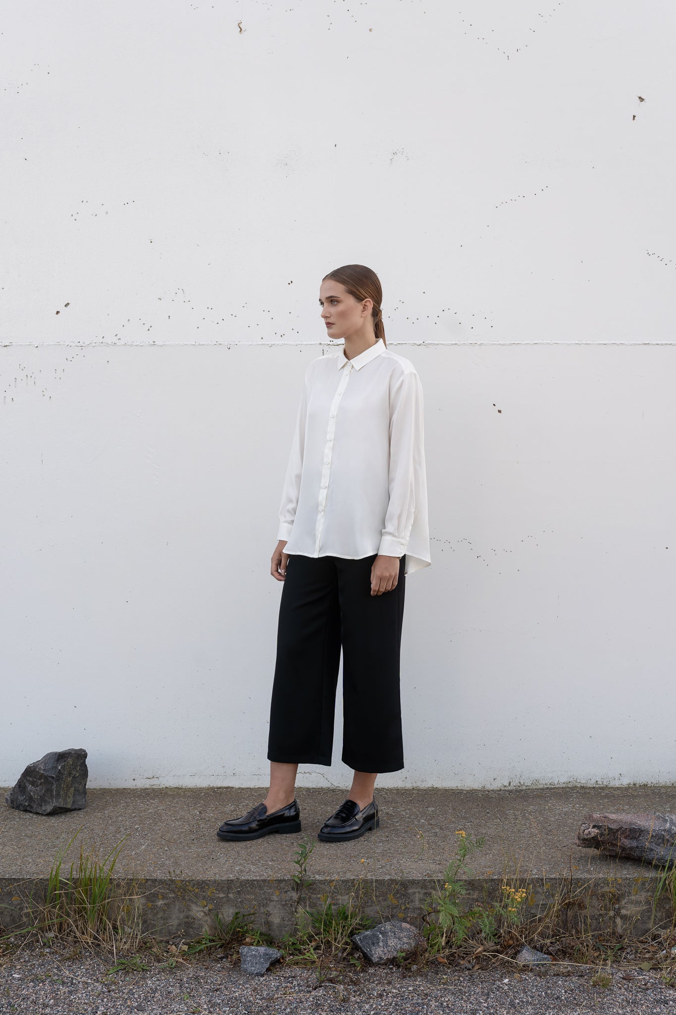 Model wearing a white silk shirt and black wool culottes standing against a plain wall.