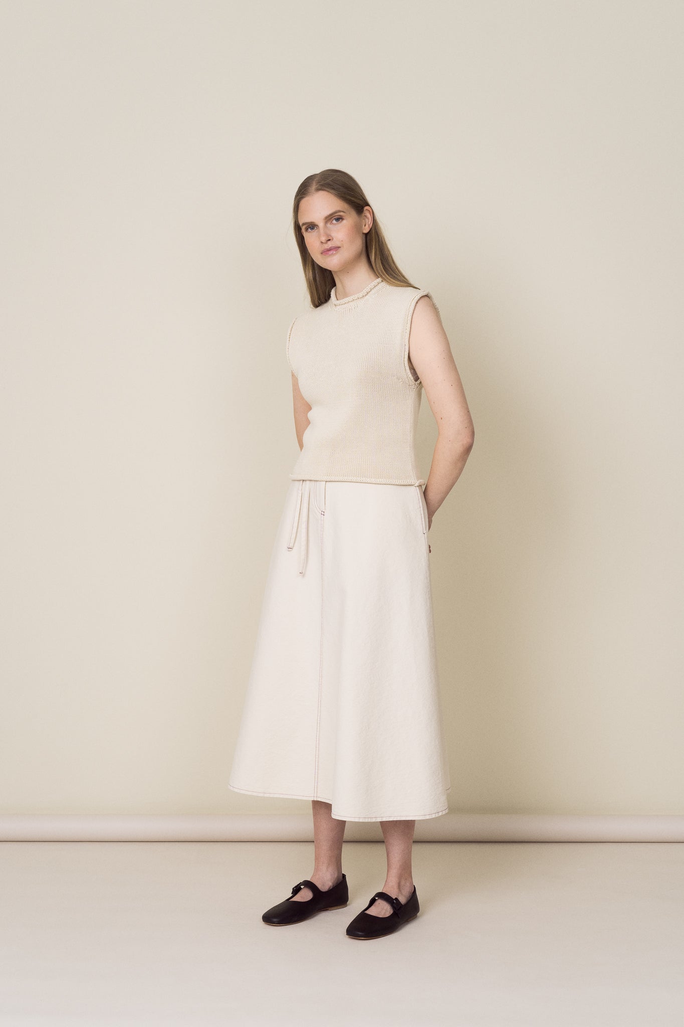 Woman wearing a sleeveless beige top and white skirt against a plain background