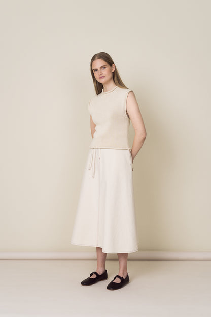 Woman wearing a sleeveless beige top and white skirt against a plain background