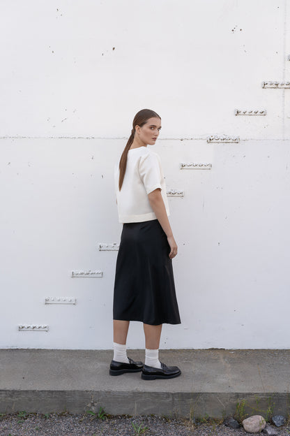 model wearing black wool midi skirt and white silk top agains industrial back wall.